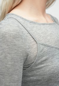 Artimų planų nuotrauka, kurioje matomas asmuo, vilkintis pilką sluoksniuotą megztinį su išryškinta medžiagos tekstūra ir iškirpte.