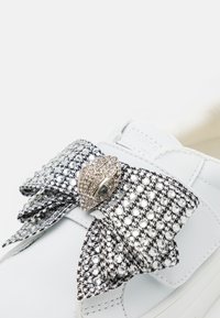 Baskets blanches avec un grand nœud noir et blanc orné de rhinestones scintillants et d'un détail métallisé.