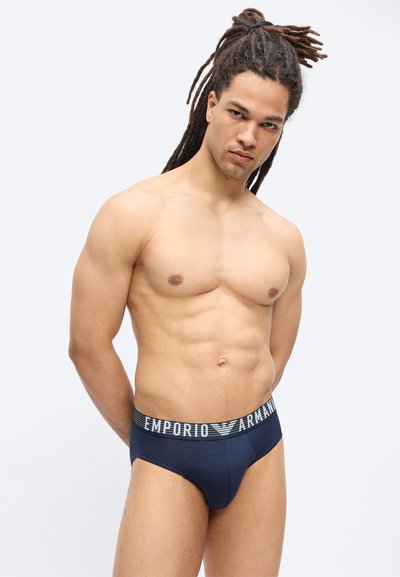 Μυώδης άνδρας με μακριά dreadlocks στέκεται μπροστά σε απλό φόντο φορώντας σκούρο μπλε εσώρουχα Emporio Armani με λογότυπο στη μέση.