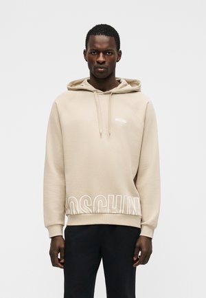 Homme noir portant un sweat à capuche beige Moschino avec des cordons de serrage et un logo blanc près de l'ourlet, assorti à un pantalon noir, debout devant un fond uni.