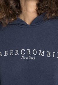 Granatowy bezrękawnik z białym haftem tekstu "ABERCROMBIE" oraz "Nowy Jork". Miękki materiał, standardowy krój i kaptur ze sznurkiem.