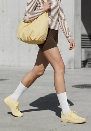 Personne marchant à l'extérieur, portant un short marron, une chemise rayée à manches longues, des chaussettes blanches, des baskets jaunes, et portant un grand sac jaune sur l'épaule.