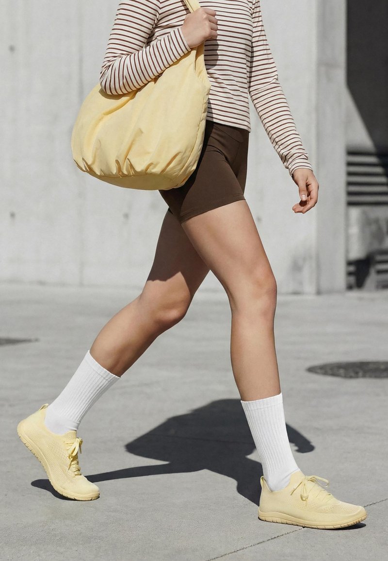Personne marchant à l'extérieur, portant un short marron, une chemise rayée à manches longues, des chaussettes blanches, des baskets jaunes, et portant un grand sac jaune sur l'épaule.
