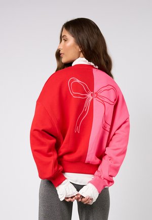 Roter und rosa Pullover mit großem Schleifenmotiv. Hergestellt aus weichem Material, mit lockerem Schnitt sowie gerippten Bündchen und Saum.