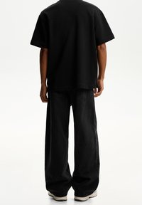 Person som bär lösa svarta jeans, svart kortärmad skjorta och vita sneakers står mot en enhetlig vit bakgrund, vänd bort.