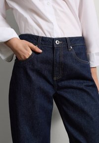Jeans de mezclilla oscura con un corte regular, costuras amarillas, cierre de botón y un bolsillo frontal. Combinados con una camisa blanca de botón.