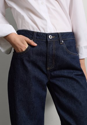 Jeans de mezclilla oscura con un corte regular, costuras amarillas, cierre de botón y un bolsillo frontal. Combinados con una camisa blanca de botón.