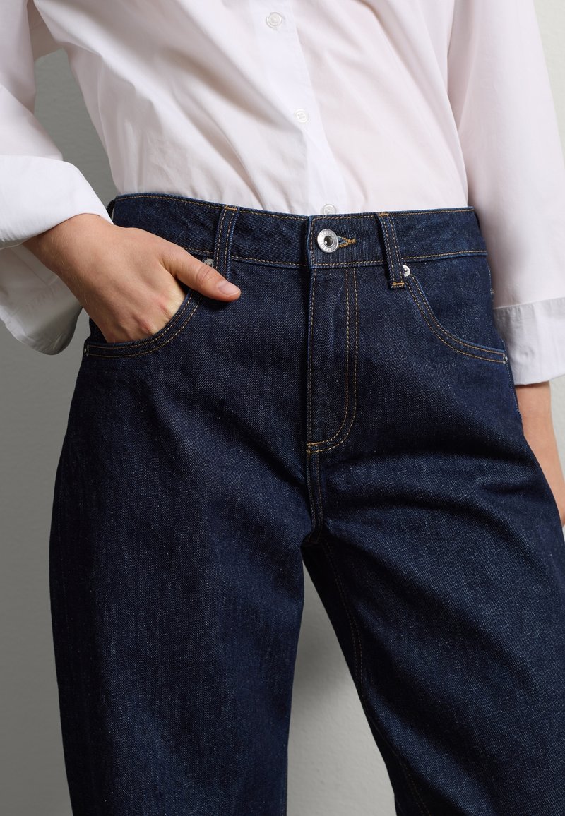 Jeans de mezclilla oscura con un corte regular, costuras amarillas, cierre de botón y un bolsillo frontal. Combinados con una camisa blanca de botón.