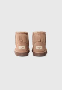 Botines cortos de ante color marrón con forro de piel de oveja, que cuentan con una suela de goma marrón plana y un detalle de costura a lo largo de las costuras. Logo de UGG visible.