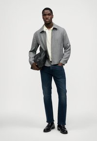 Jaqueta cinza com zíper sobre uma polo bege, combinada com jeans escuros e sapatos pretos. O modelo segura uma bolsa preta texturizada. Design simples e limpo.