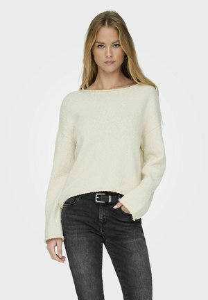 Pull léger crème à col bateau large et manches longues, avec un tricot texturé. Associé à un jean en denim foncé et une ceinture noire.