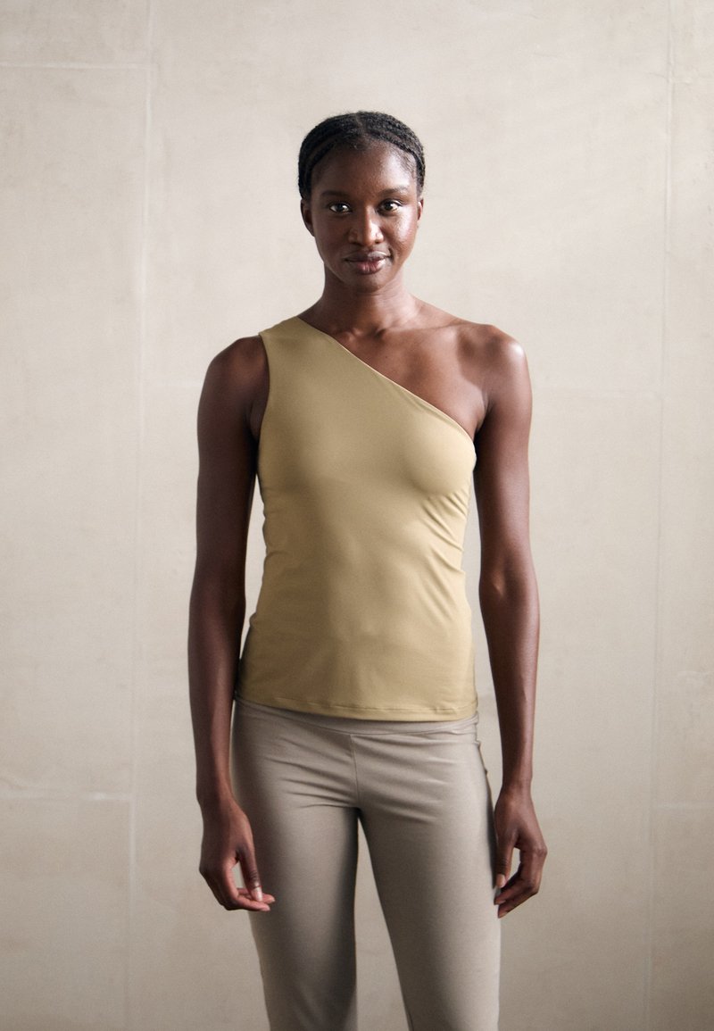 Top a spalla asimmetrico in tessuto beige, texture liscia, design aderente, abbinato a leggings beige chiari.