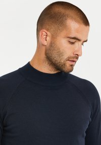 Marineblauer Strickpullover mit hohem Kragen, gerippter Textur und Raglanärmeln, der eine taillierte Form betont.