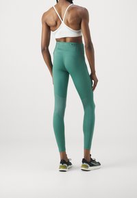 Vrouw in petrolkleurige hoge taille legging en een witte sportbeha met bandjes, staand met haar rug naar de camera tegen een effen witte achtergrond.