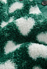 Tessuto in peluche verde e bianco con un design testurizzato caratterizzato da cuori soffici. Lo sfondo è di un verde intenso, che enfatizza il contrasto con il bianco.