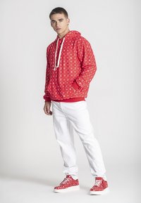 Joven con sudadera roja estampada y zapatillas a juego, vestido con pantalones blancos, de pie frente a un fondo gris claro liso.