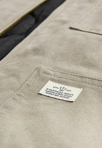 Beige kangasta, jonka pinnassa on tekstuuria, ja siinä on valkoinen etiketti, jossa lukee "STANDARD ISSUE", sekä vetoketjuhauki.
