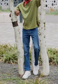 T-shirt en coton vert, jeans en denim bleu, baskets blanches, bandana rouge. Posé entre de fins troncs d'arbres blancs avec de l'herbe et du gravier sous les pieds.