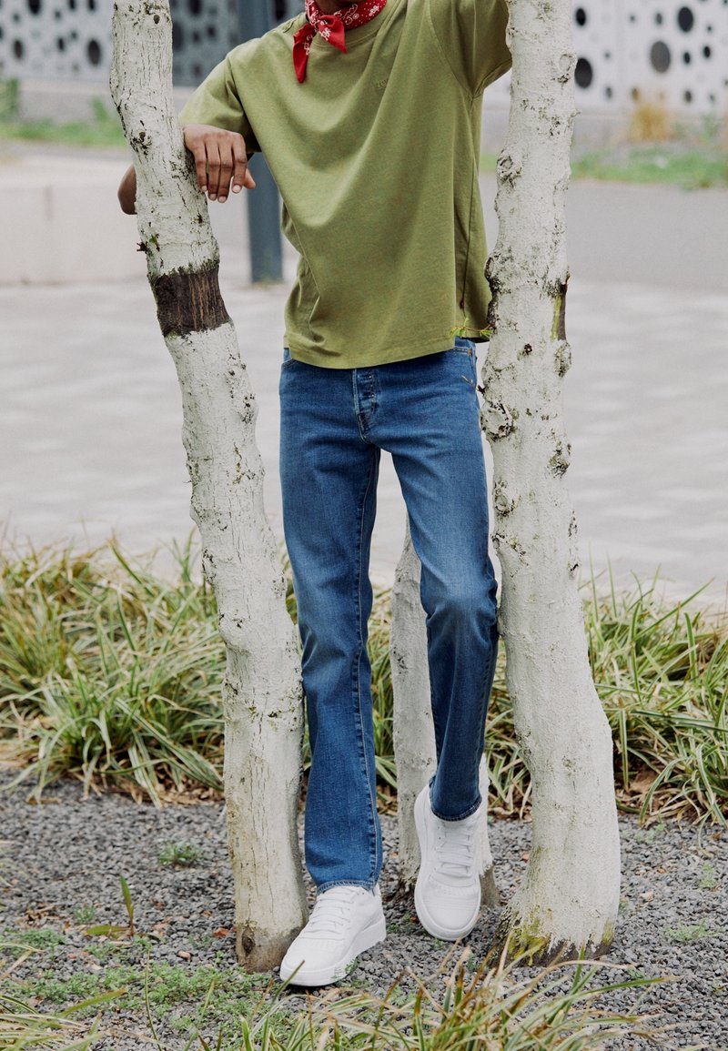 T-shirt en coton vert, jeans en denim bleu, baskets blanches, bandana rouge. Posé entre de fins troncs d'arbres blancs avec de l'herbe et du gravier sous les pieds.