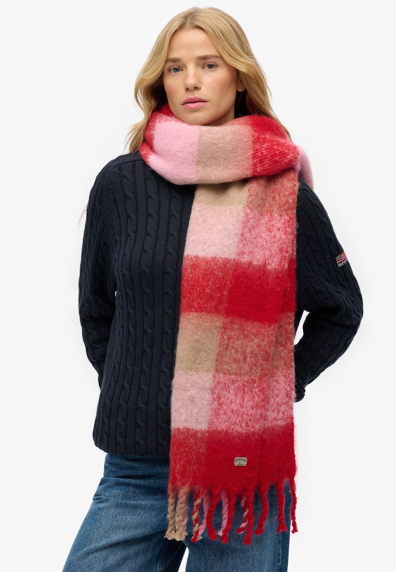 Uldhalstørklæde i rød, pink og beige ternet mønster med frynser i kanten; blød tekstur der hænger omkring halsen, båret med en navy kabelstrikket sweater.