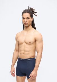 Μυώδης άνδρας με κοτσίδες dreadlocks που φοράει μαύρα μποξεράκια Calvin Klein, στέκεται μπροστά σε απλό λευκό φόντο.