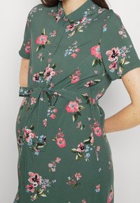 Abito verde floreale con maniche corte, chiusura a bottoni frontale, cintura in vita, con fiori rosa e blu e un silhouette rilassata.