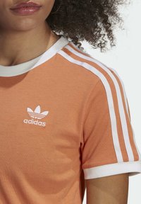 Orange bomullströja med vit ribbad krage, vita tre ränder på axlarna och broderad vit Adidas-logga på bröstet.