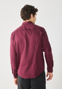 Burgunderfarbene Langarmbluse mit glatter Textur, Button-Down-Kragen und abgerundetem Saum. Verfügt über Manschetten mit Knopfverschluss.