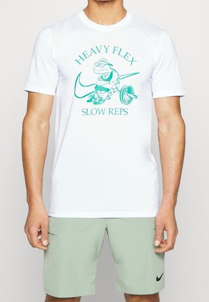 Mann trägt weißes T-Shirt mit grünem Motiv einer muskelnden Schildkröte und dem Text "Heavy Flex Slow Reps" mit einer Langhantel- und Schwung-Logo.