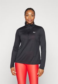 Haut de sport à manches longues noir avec un col zippé et un détail de logo blanc. Le matériau est lisse et ajusté, associé à des leggings rouges.