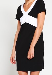Kobieta w czarnej sukience z krótkimi rękawami i białym wzorem w kształcie litery V na klatce piersiowej, stojąca na jednolitym jasnym tle.