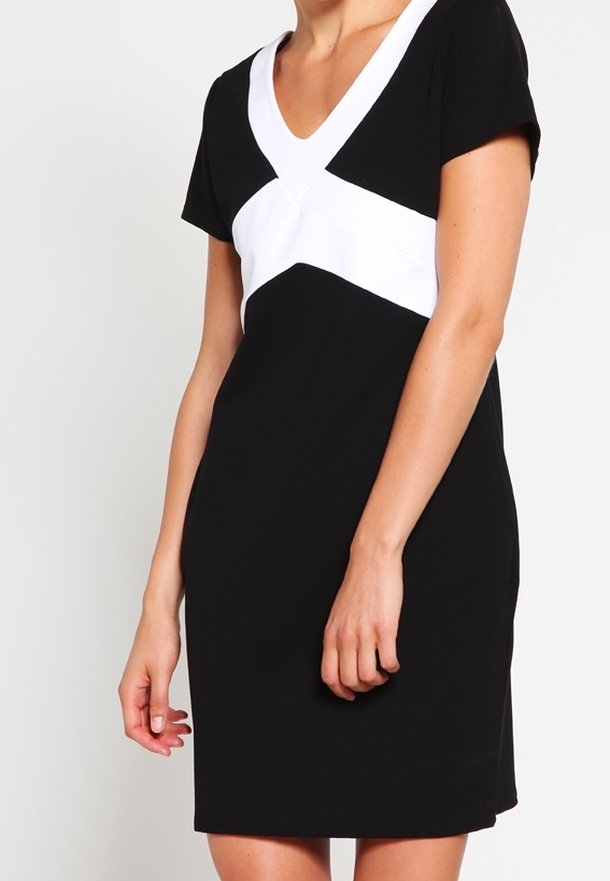 Kobieta w czarnej sukience z krótkimi rękawami i białym wzorem w kształcie litery V na klatce piersiowej, stojąca na jednolitym jasnym tle.