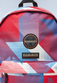 Mochila de tela con patrón geométrico en rojo y azul, con un parche de logo redondo y una etiqueta de marca rectangular en la parte frontal. Acentos en negro.