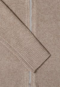 Beige stickad cardigan med ribbat mönster, med en silverfärgad dragkedja framtill och långa, avsmalnande ärmar. Mjuk, ull-liknande material.