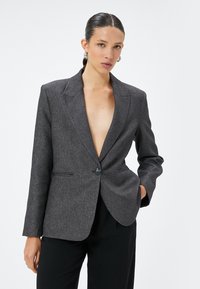 Grijze op maat gemaakte blazer met notch-lapels, enkele knoopsluiting en twee voorklauwen; de textuur van de stof biedt een subtiel patroon.