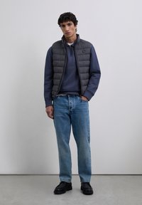 Joven vestido con un chaleco acolchado negro sobre un suéter azul marino de media cremallera, jeans azul claro y zapatos negros, de pie contra un fondo sencillo.