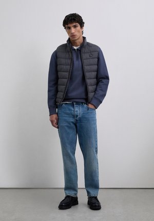 Junger Mann, der eine schwarze gesteppte Weste über einem marineblauen Halbzip-Pullover trägt, dazu hellblaue Jeans und schwarze Schuhe, stehend vor einem schlichten Hintergrund.