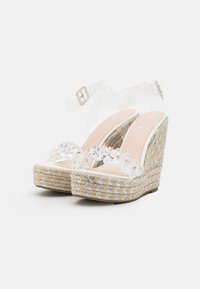 Sandalias de cuña transparentes con una base de yute trenzado, adornadas con correas transparentes y piedras iridiscentes en la parte frontal. Diseño elegante y moderno.