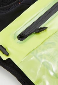 Tessuto nero con una tasca con cerniera verde lucido; presenta accessori neri e una superficie testurizzata. Include un cordino per una facile apertura.