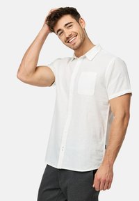 Camisa de lino blanca de manga corta con cuello abotonado y bolsillo en el pecho izquierdo. Presenta un ajuste relajado y una tela texturada.