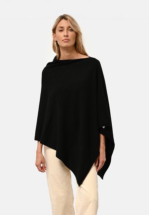 Svart poncho med vid halsringning, asymmetrisk fåll och mjuk textur, i kombination med ljusa byxor.