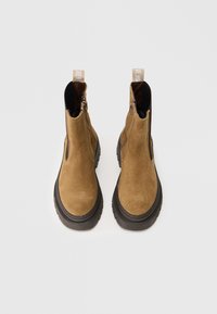 Botines de ante marrón con punta redonda, cremalleras laterales y una suela de goma negra gruesa. Incluyen tiradores para facilitar su uso.