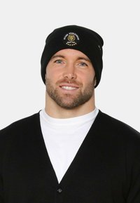 Uomo con barba che indossa un berretto nero con il logo dello Stade Toulousain, una maglietta bianca a girocollo e un cardigan nero, sorridente su uno sfondo chiaro.