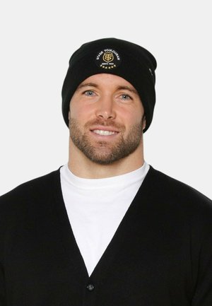 Hombre con barba que lleva un gorro negro con el logo del Stade Toulousain, camiseta blanca de cuello barco y cárdigan negro, sonriendo frente a un fondo claro.