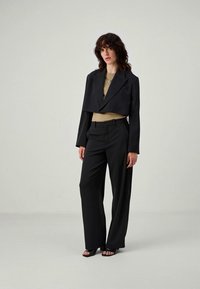 Schwarze, kurz geschnittene Blazerjacke, kombiniert mit hochgeschnittenen schwarzen Hosen. Das Outfit umfasst ein beiges Oberteil darunter und schwarze, schnürbare High Heels.
