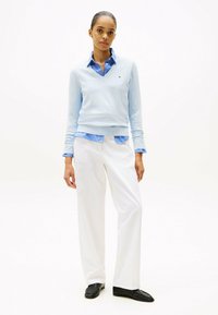 Pull azzurro chiaro sovrapposto a una camicia a righe blu, abbinato a pantaloni bianchi e mocassini neri. I tessuti sono lisci e sagomati.