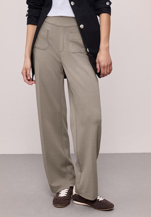 Personne portant un pantalon beige à jambes larges avec poches plaquées à l'avant, une longue veste noire à boutons, une chemise blanche et des baskets marron foncé avec des lacets blancs.