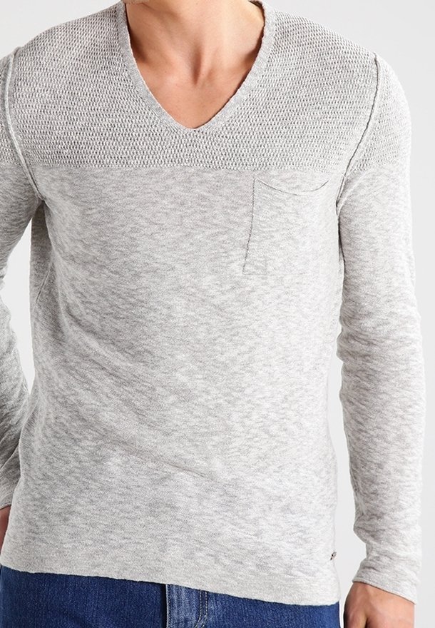 Personne portant un pull en maille texturée gris clair à col en V, petite poche poitrine et jean bleu, montrée des épaules aux hanches.