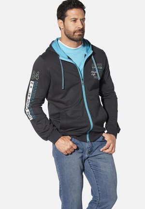 Black zip-up hoodie with blue accents, featuring a drawstring hood, printed text on the sleeves, and ribbed cuffs, paired with blue jeans.