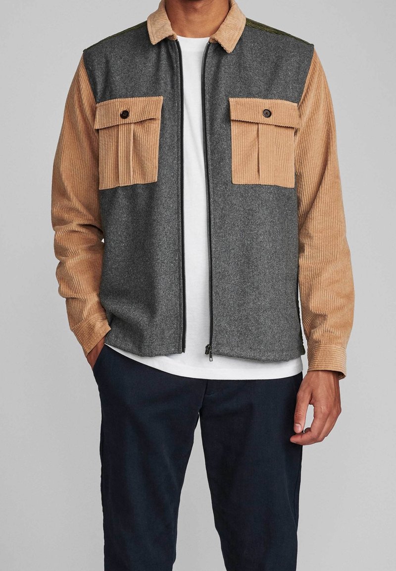 Homme portant une veste à blocs de couleurs avec un corps gris et des manches et poches poitrine en velours côtelé beige, chemise blanche et pantalon sombre.
